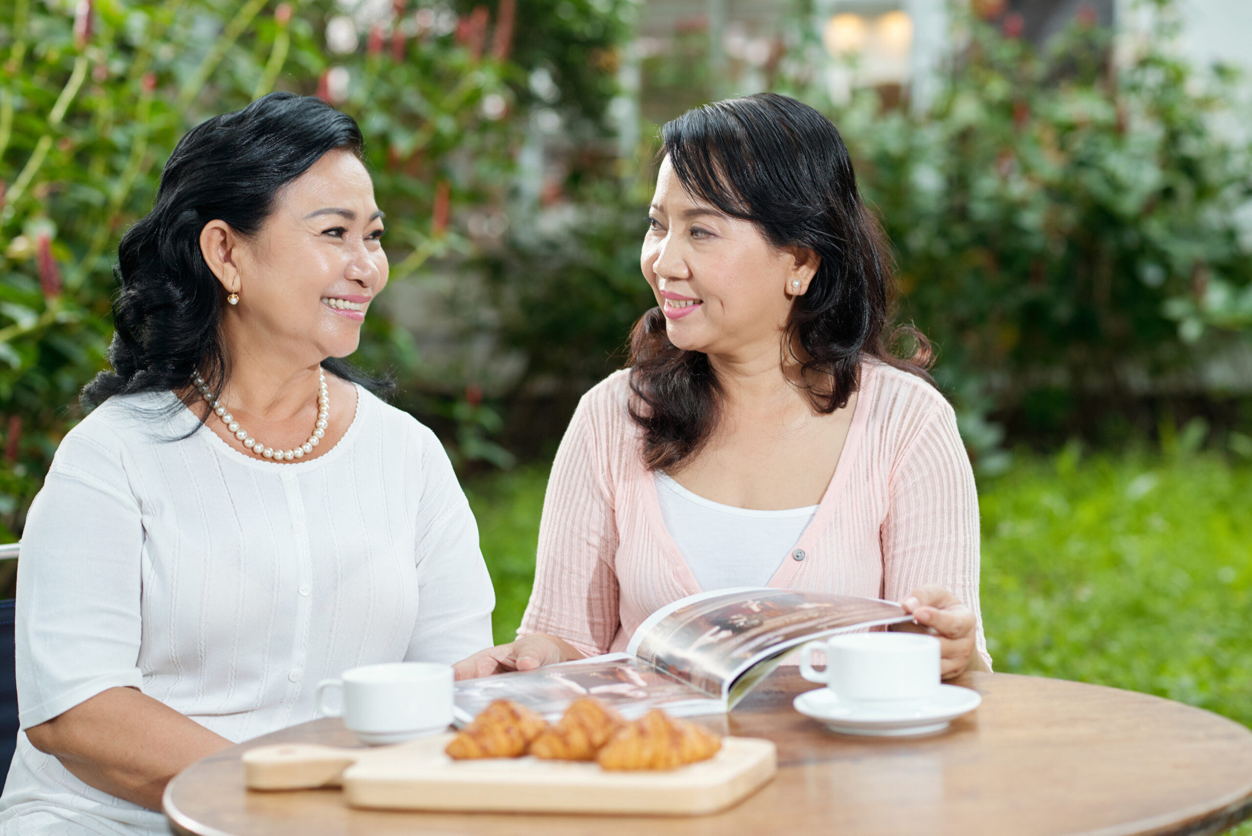 Attractive stylish aged Asian women chatting and smiling cheerfully while sitting at table outdoors with tea and croissants