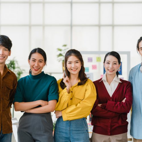 Group of Asia young creative people in smart casual wear smiling and arms crossed in creative office workplace. Diverse Asian male and female stand together at startup. Coworker teamwork concept.
